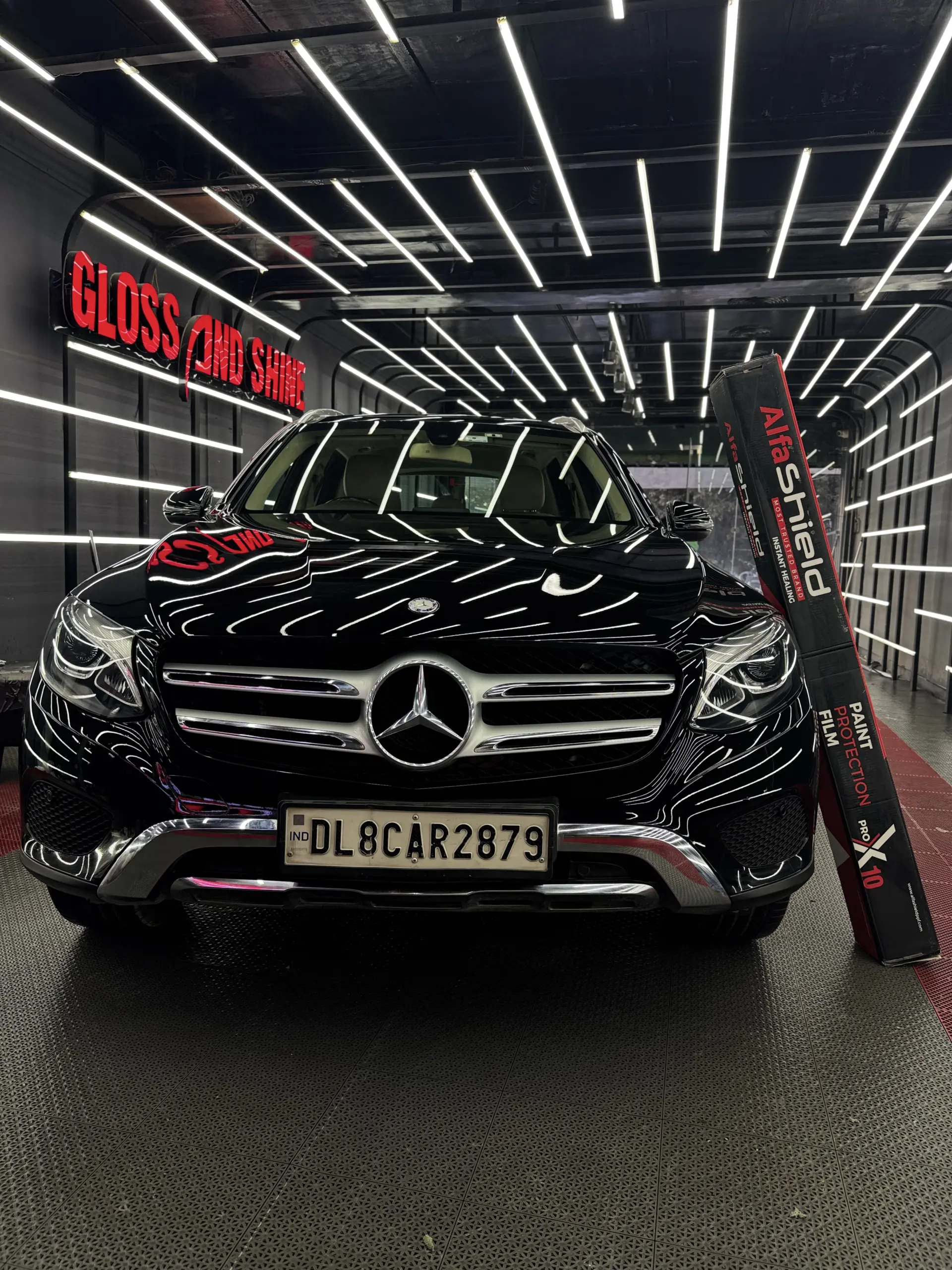 Black Mercedes-Benz SUV in a premium detailing studio with LED ceiling lights, showcasing AlfaShield Paint Protection Film box, highlighting high-gloss finish, car protection, and luxury detailing environment.