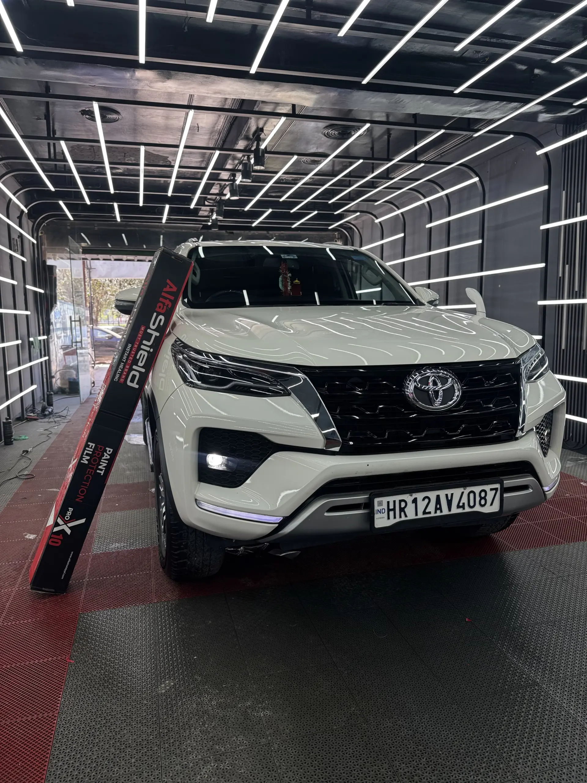 White Toyota Fortuner in a premium car detailing studio with LED lights, showcasing AlfaShield Paint Protection Film box, highlighting high gloss finish and advanced car protection.