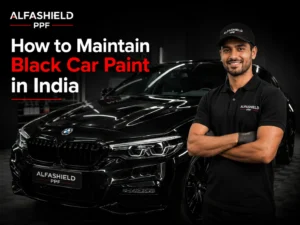A professional promotional banner featuring a glossy black luxury car inside a dark detailing studio with a smiling man wearing a black Alfashield PPF t-shirt and cap. The image includes the title “How to Maintain Black Car Paint in India” in bold white and red text, along with Alfashield PPF branding and dramatic black shadow lighting for a premium automotive look.